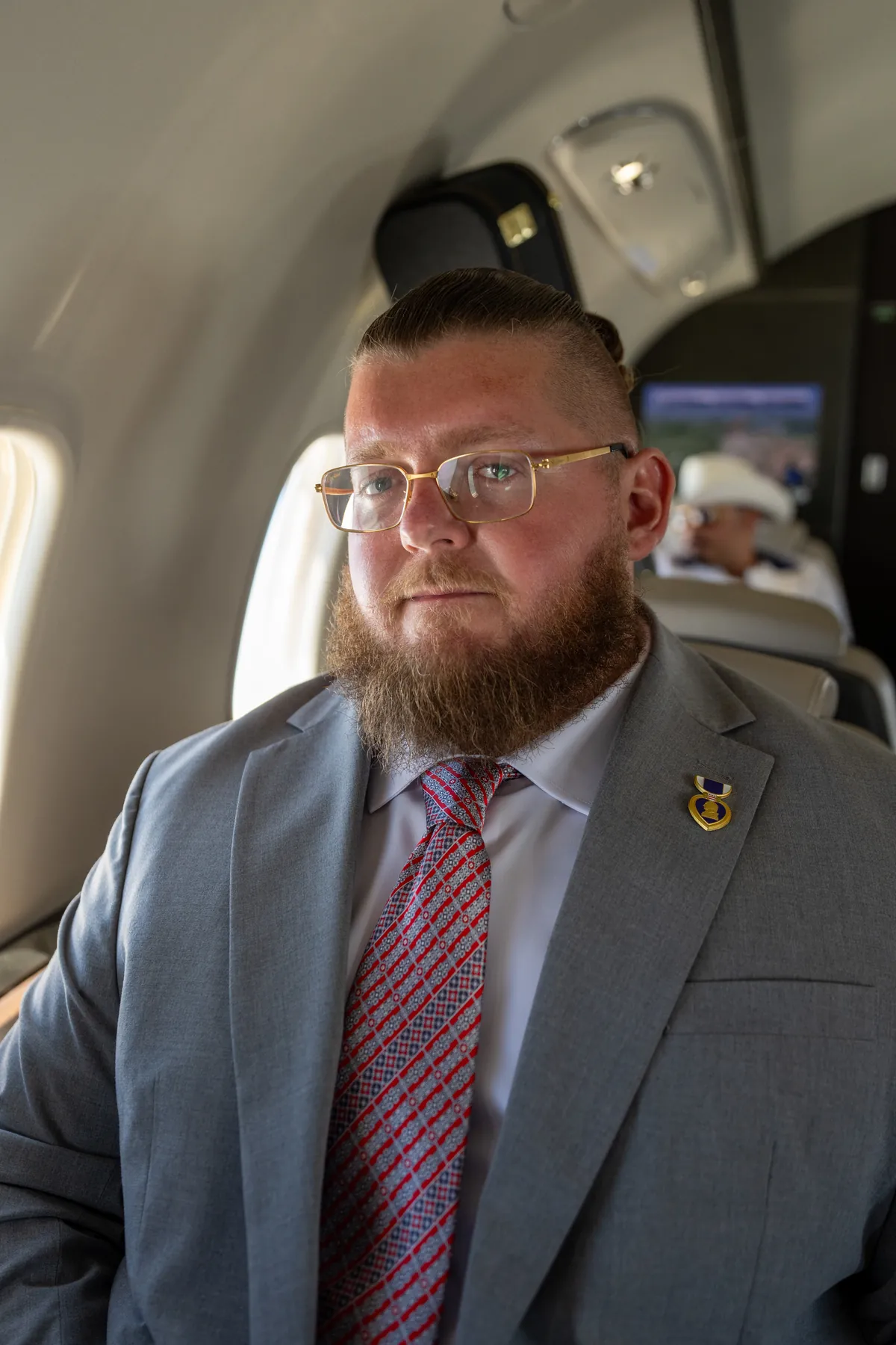 Lukas Simianer in a grey suit and red tie seated in an aircraft cabin, looking off camera past the window.